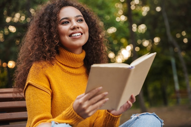 Retrato de mujer rizada hermosa complacida alegre sentarse en el parque al aire libre leyendo el libro.