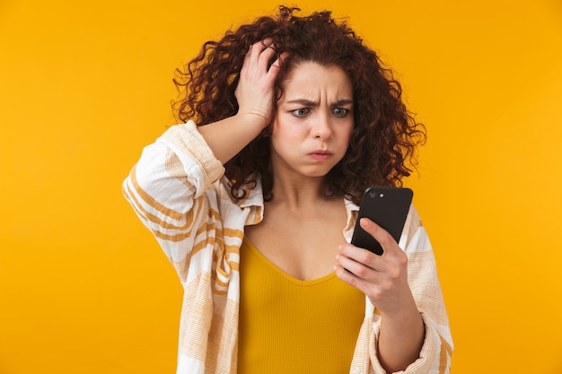Foto retrato de una mujer rizada joven confusa disgustada hermosa linda que presenta aislada en la pared amarilla que usa el teléfono móvil.