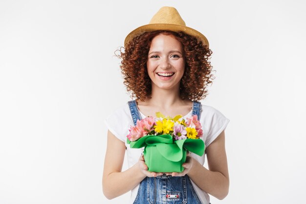 Retrato de mujer rizada pelirroja femenina de 20 años con sombrero de paja de verano sonriendo y sosteniendo la caja de flores aislada sobre la pared blanca