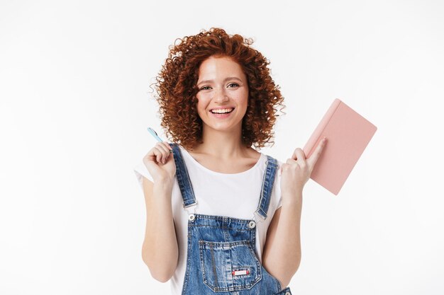 Retrato de mujer rizada pelirroja positiva de 20 años vistiendo mono de mezclilla sonriendo y escribiendo notas en su diario o planificador diario aislado sobre pared blanca