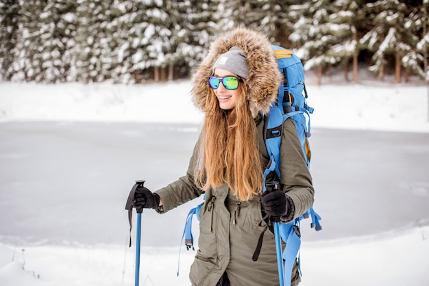 Retrato de una mujer en ropa de invierno de senderismo con mochila y palos de rastreo en el bosque nevado cerca del lago congelado