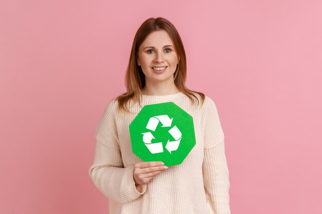 Retrato de una mujer rubia adulta joven positiva feliz sosteniendo un cartel de reciclaje verde salvando el concepto de ecología ambiental usando un suéter blanco Foto de estudio interior aislada sobre fondo rosa