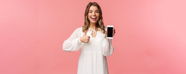 Retrato de mujer rubia alegre y guapa vestida de blanco recomienda descargar la aplicación o suscribirse usando la aplicación de compras en línea hacer que el pulgar muestre la pantalla del teléfono móvil