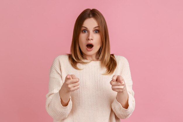 Retrato de mujer rubia apuntando con el dedo a la cámara, luciendo sorprendida con la boca abierta, difícil de creer, usando suéter blanco. Disparo de estudio interior aislado sobre fondo rosa.