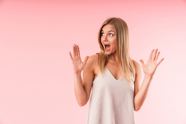 Retrato de mujer rubia asombrada expresando asombro y levantando las manos mientras mira a un lado en copyspace aislado sobre pared rosa en estudio