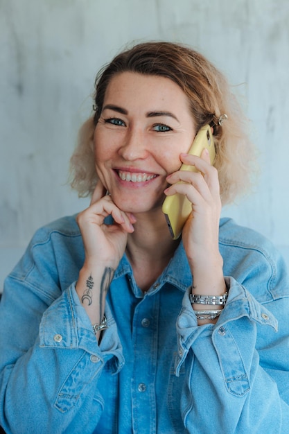 Retrato de una mujer rubia complacida con una camisa de mezclilla casual hablando por un teléfono móvil en una casa moderna