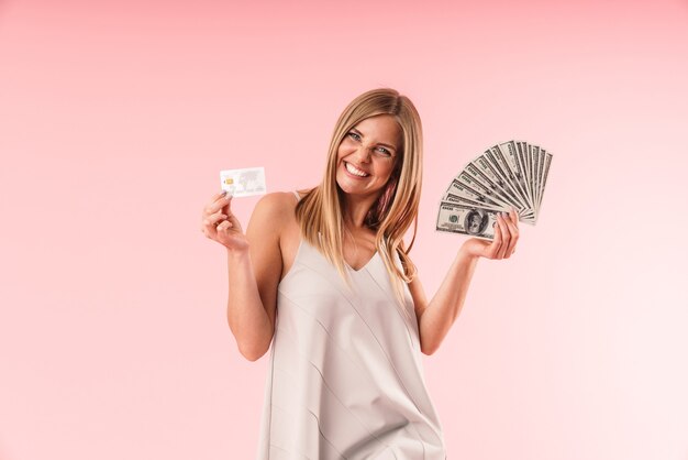 Retrato de mujer rubia emocionada sonriendo mientras sostiene tarjetas de crédito y facturas de dinero aisladas sobre pared rosa en estudio