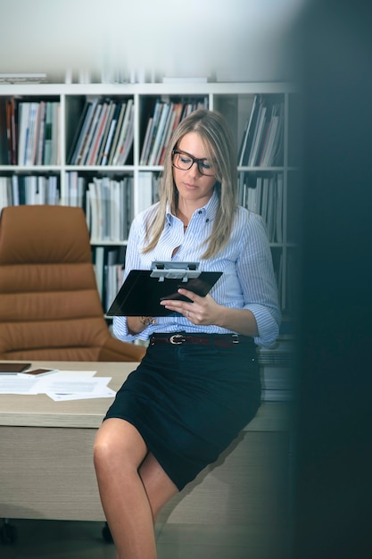 Retrato de mujer rubia escribiendo notas sentado sobre su mesa de escritorio. Ver el canal de la puerta de la oficina.