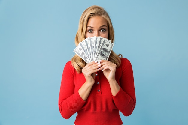 Retrato de mujer rubia europea 20s vistiendo camisa roja sosteniendo ventilador de dinero en dólares, aislado sobre pared azul