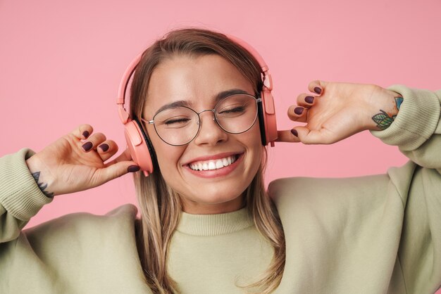 Retrato de mujer rubia feliz en anteojos usando auriculares y bailando aislado sobre pared rosa