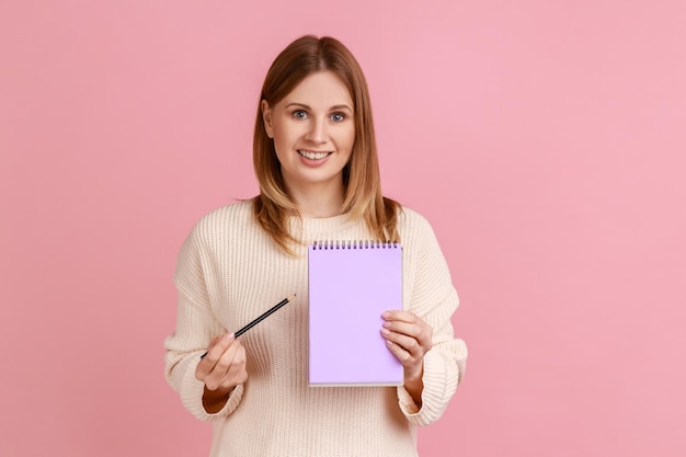 Retrato de una mujer rubia feliz y satisfecha señalando un cuaderno de papel mirando sonriendo a la cámara tiene una expresión positiva usando suéter blanco Foto de estudio interior aislada en fondo rosa