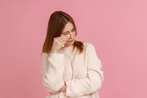 Retrato de mujer rubia frunciendo el ceño, limpiando lágrimas llorando, sintiéndose desesperada sin esperanza, lidiando sola con estrés emocional, usando suéter blanco. Disparo de estudio interior aislado sobre fondo rosa.