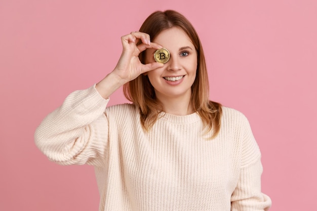 Retrato de una mujer rubia graciosa que cubre el ojo con tecnología de cadena de bloques de bitcoin e inversión electrónica de criptomonedas con suéter blanco Foto de estudio interior aislada en fondo rosa