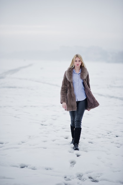 Retrato de mujer rubia joven elegancia en un abrigo de piel, río brumoso en hielo de invierno.