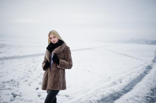 Retrato de mujer rubia joven elegancia en un abrigo de piel, río brumoso en hielo de invierno.