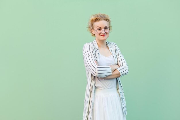 Foto retrato de mujer rubia joven feliz en camisa blanca, falda y blusa a rayas con anteojos de pie, brazos cruzados y mirando a cámara con una sonrisa. tiro del estudio aislado en fondo verde claro.