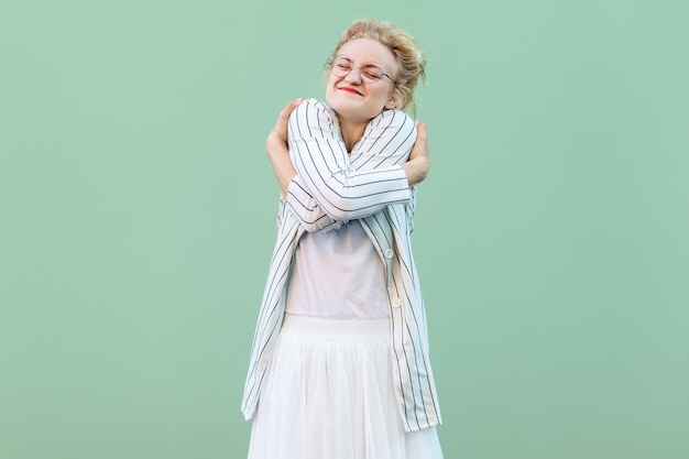 Retrato de mujer rubia joven orgullosa feliz con camisa blanca, falda y blusa a rayas con anteojos de pie abrazándose a sí misma y sintiéndose bien. tiro del estudio de interior aislado en fondo verde claro.