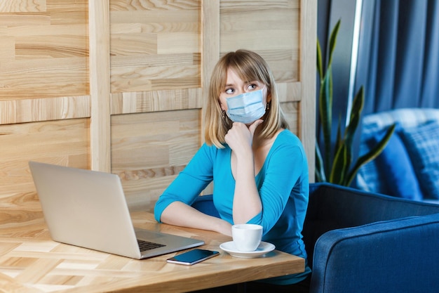 Retrato de mujer rubia joven pensativa con máscara médica quirúrgica en camiseta azul está sentado y trabajando en la computadora portátil y pensando en su trabajo. Concepto de trabajo, medicina y salud en interiores.