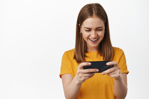 Retrato de mujer rubia jugando juegos de video en el teléfono móvil. Mujer joven viendo videos en internet celular de teléfonos inteligentes, sonriendo y riendo, mire la pantalla, pared blanca.