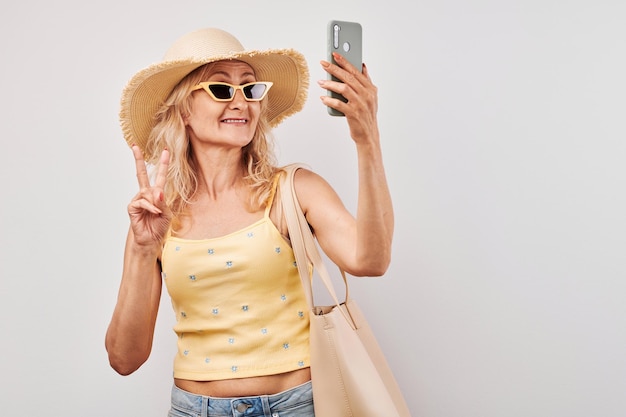 Retrato mujer rubia madura positiva en sombrero de paja top amarillo y gafas de sol sosteniendo teléfono inteligente y bolsa de compras aislado sobre fondo blanco de estudio