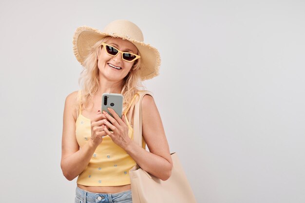 Retrato mujer rubia madura positiva en sombrero de paja top amarillo y gafas de sol sosteniendo teléfono inteligente y bolsa de compras aislado sobre fondo blanco de estudio