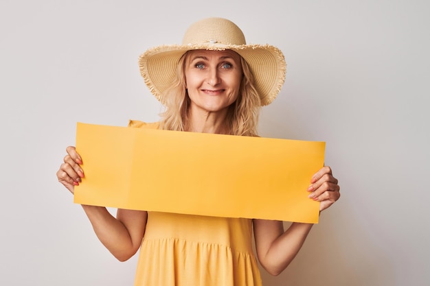 Retrato mujer rubia madura positiva en sombrero de paja y vestido amarillo sosteniendo cartel amarillo en blanco en las manos con espacio de copia de texto aislado sobre fondo blanco de estudio