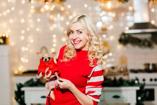 Retrato de mujer rubia con Navidad Santa con perros chihuahua en traje de Navidad en la cocina con decoración navideña, sonriendo y mirando a cámara.