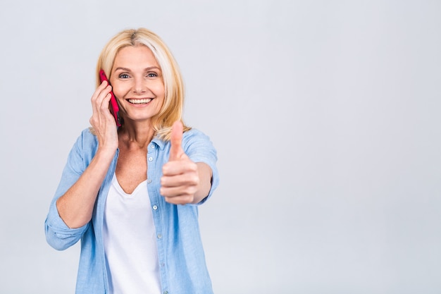 Retrato de mujer rubia de negocios senior madura hermosa alegre mediante teléfono móvil. Sonriendo, aislado sobre fondo gris blanco. Pulgar hacia arriba signo.