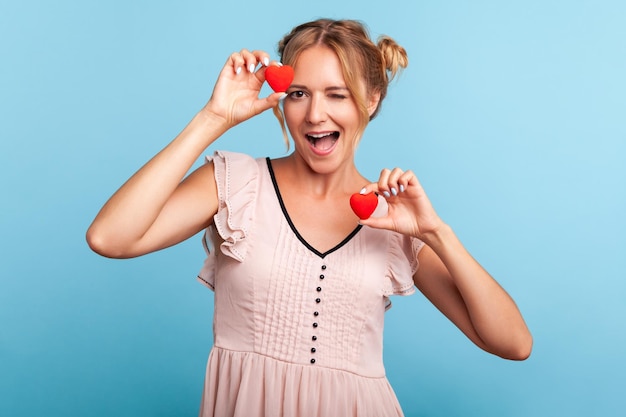 Retrato de una mujer rubia positiva y amistosa vestida con dos bollos de pelo sosteniendo símbolos de corazón en las manos y guiñando un ojo a la cámara enviando amor Disparo de estudio interior aislado sobre fondo azul