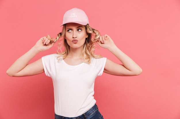 Foto retrato de mujer rubia rizada con camiseta casual y gorra sonriendo al frente aislado sobre pared rosa