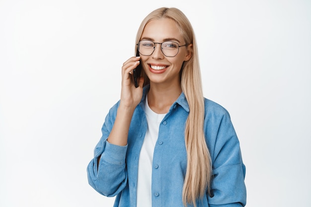 Retrato de mujer rubia sonriente con gafas hablando por teléfono móvil, con una llamada de teléfono inteligente, de pie en blanco.