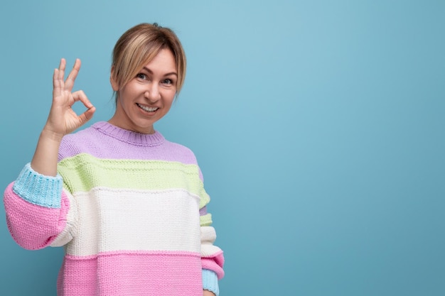 Retrato de una mujer rubia sonriente con ropa informal que muestra un gesto correcto en el fondo azul con copia