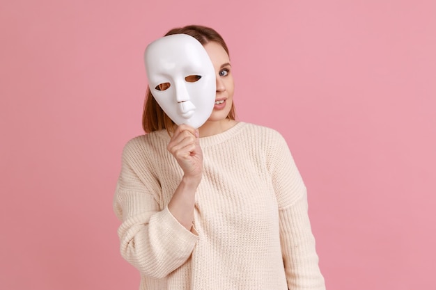 Retrato de una mujer rubia sonriente sosteniendo una máscara blanca mirando a la cámara fingiendo ser otra persona con suéter blanco Foto de estudio interior aislada en fondo rosa