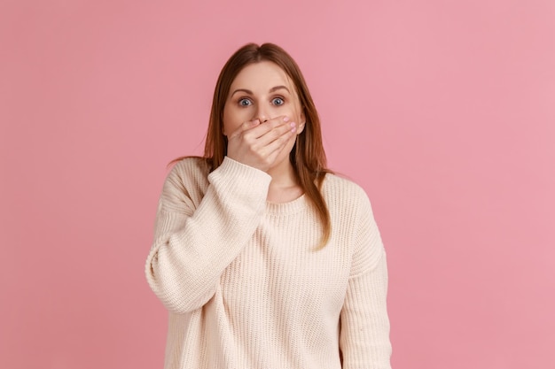 Retrato de una mujer rubia sorprendida que se puso la mano en la boca y miró con miedo a los ojos manteniendo un terrible secreto usando un suéter blanco Foto de estudio interior aislada en un fondo rosa