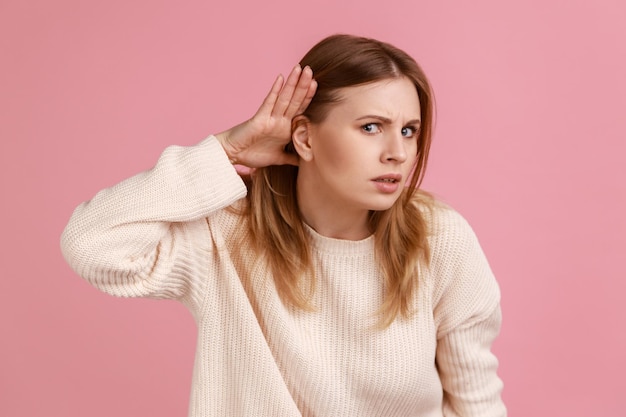 Foto retrato de mujer rubia sosteniendo la mano cerca de la oreja y escuchando atentamente, con problemas de audición, sordera en la comunicación, usando suéter blanco. disparo de estudio interior aislado sobre fondo rosa.