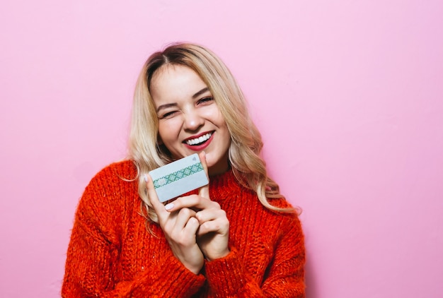 Retrato de una mujer rubia con una tarjeta de crédito y sonriendo en la habitación en una pared rosa