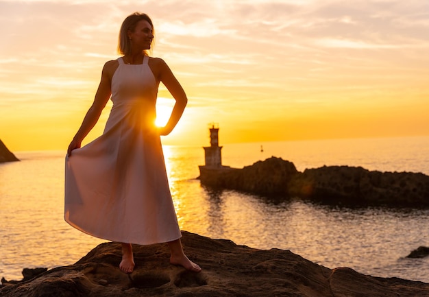 Retrato de una mujer rubia con un vestido blanco al atardecer junto a un faro en el mar estilo de vida saludable y naturista con el sol de fondo