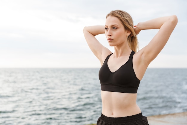 Retrato de mujer seductora linda vistiendo chándal haciendo ejercicio mientras se ejercita en el muelle cerca de la playa en la mañana