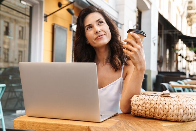 Retrato de mujer seductora soñando con bolsa de paja bebiendo café de la taza de papel y usando la computadora portátil mientras está sentado en la acogedora cafetería al aire libre