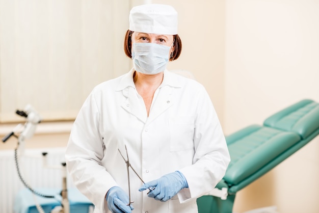 Retrato de una mujer senior ginecóloga en bata médica, gorro y guantes de pie con pinzas en la oficina ginecológica