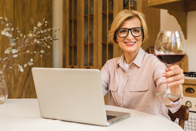 Retrato de mujer senior madura sonriente sosteniendo una copa de vino mientras usa el portátil en la mesa de la cocina. Freelance trabajando en concepto de hogar.