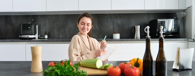 Retrato de una mujer sentada con comestibles haciendo una lista para ir de compras posando cerca de preparar verduras