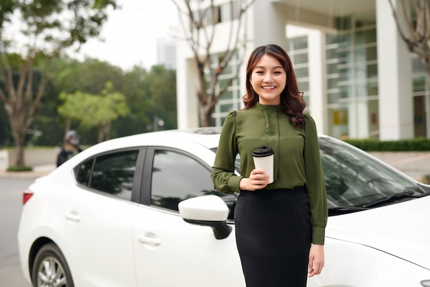 Retrato de mujer sexy sonriente bebiendo taza de bebida mientras está parado cerca del coche afuera