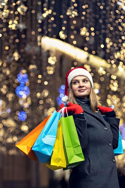 Foto retrato de una mujer con un sombrero de santa con compras navideñas en bolsas de colores en la calle decoradas