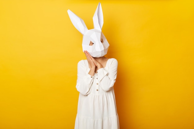 Retrato de una mujer somnolienta con vestido blanco y máscara de conejo de papel parada aislada sobre fondo amarillo necesita tomar una siesta mantiene las palmas cerca de su cara dormida