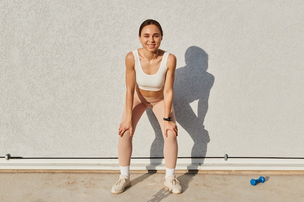 Retrato de mujer sonriente adulta joven vistiendo top deportivo blanco y leggins beige de pie con las manos en la rodilla, descansar después de correr, respirar, mirando a la cámara.