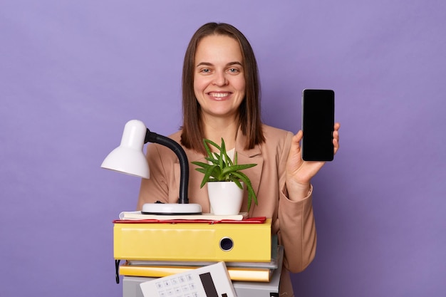 Retrato de una mujer sonriente y atractiva con una chaqueta beige que sostiene muchas carpetas de documentos que muestran un teléfono móvil con un espacio vacío para la promoción aislado sobre un fondo lila
