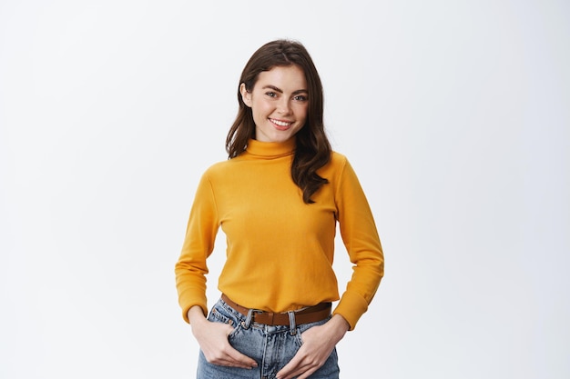 Foto retrato de mujer sonriente atractiva con rostro natural sano con maquillaje ligero, tomados de la mano en los bolsillos de los pantalones vaqueros y mirando confiado en blanco