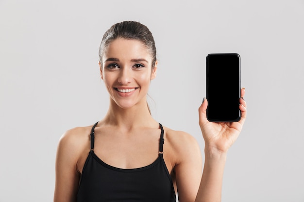 Retrato de mujer sonriente bien construida que muestra la pantalla negra del teléfono, aislado sobre la pared gris