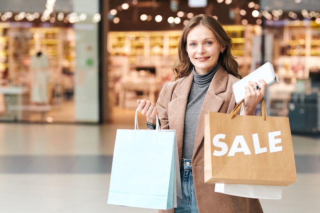 Retrato de mujer sonriente con bolsas de compras y bolso disfrutando de la venta de temporada en el centro comercial